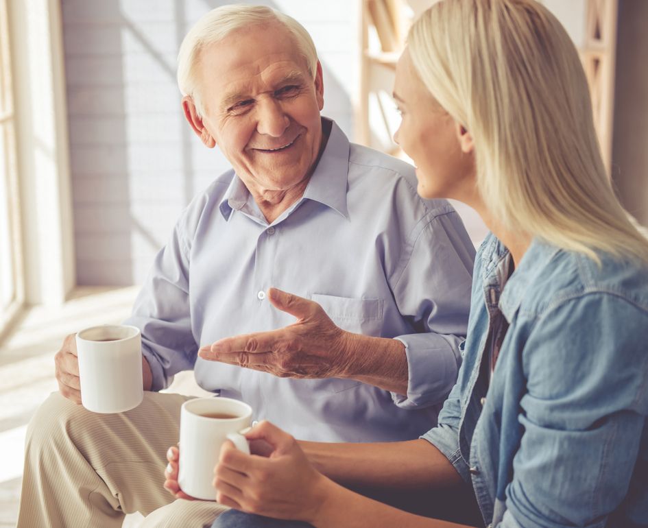 Auf einer Couch sitzen ein älterer Mann und eine ältere Frau, beide halten Kaffeetassen und wirken entspannt und zufrieden.